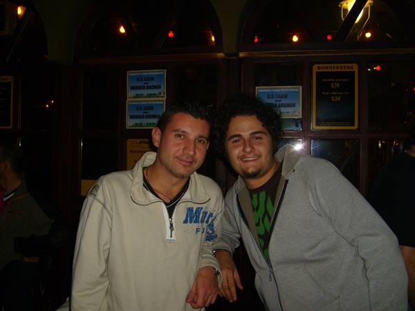 Two friends smile in front of a dark wooden pub bar, soft lights