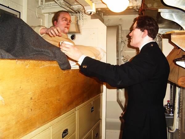 Wax figures in a ship cabin: sailor in bunk, officer handing a cup