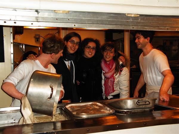 Smiling cooks serve fried food from a steel counter, tourists posing alongside