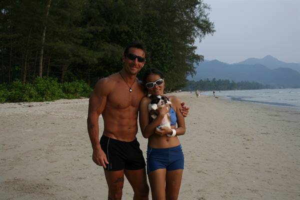 Couple in swimwear on sandy beach, she hugs a puppy, pines and mountains