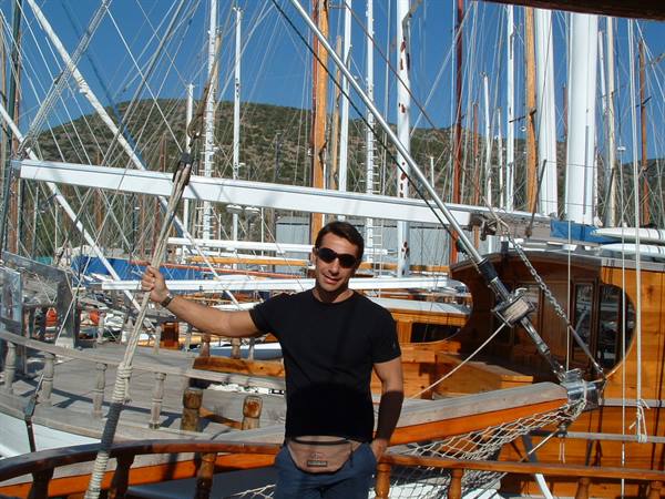 Man in black t-shirt on wooden gulet among boat masts, sunny light