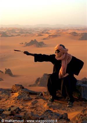 tuareg man in turban points at sandy dunes among desert rocks