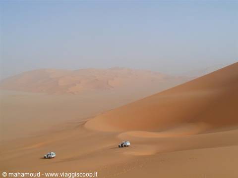 Two white jeeps climbing huge orange sand dunes under hazy sky
