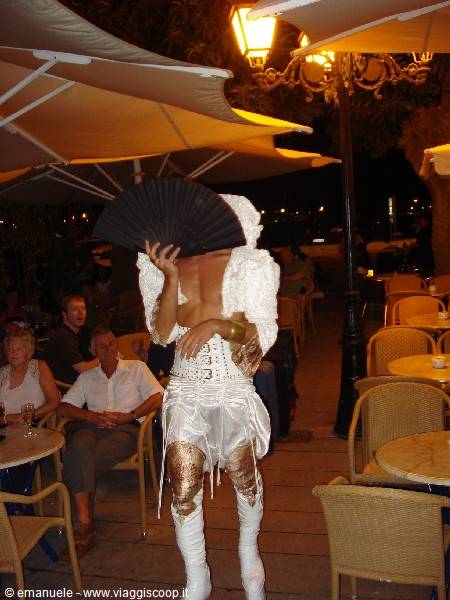 Drag queen in white and gold with black fan among night café tables