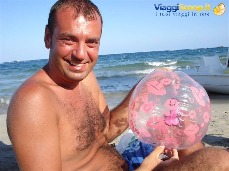 Smiling man on beach holding inflated pink ball, sea waves and white boat