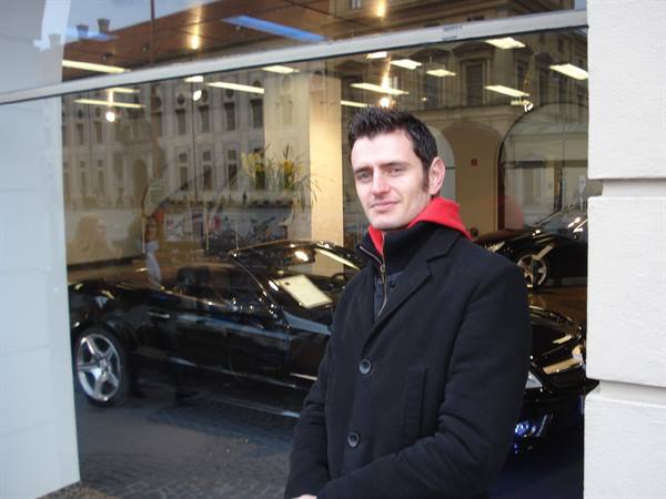 Man in black coat with red hood outside a Mercedes dealership showroom window