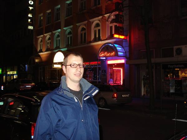 Man in glasses and blue jacket in front of hotel lit by neon signs at night