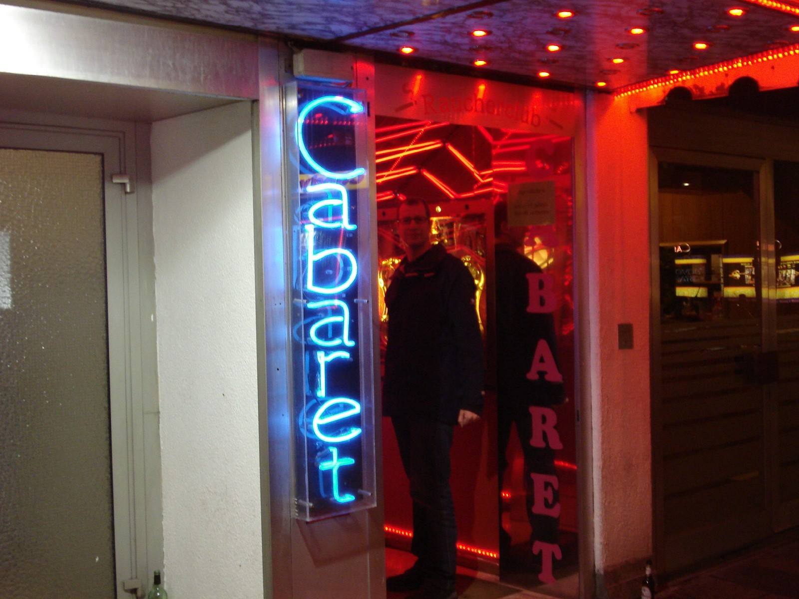 Man standing at a cabaret entrance, blue and red neon signs at night