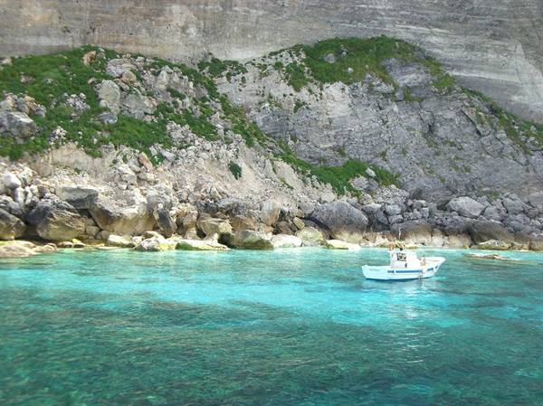 White boat on clear turquoise water beneath rocky cliffs with green shrubs