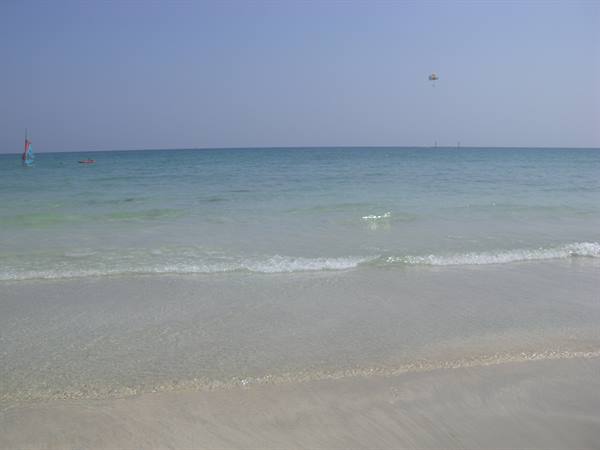 Gentle waves on pale sand, windsurf sail left, parasailing in the sky