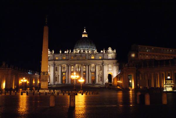 St. Peter's Square at night: illuminated basilica, obelisk, lights reflecting on wet cobblestones