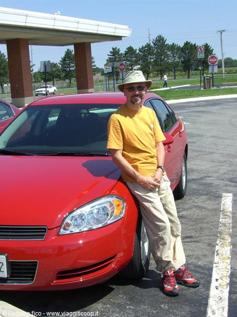 Man in hat and yellow shirt leaning against a red car in a parking lot