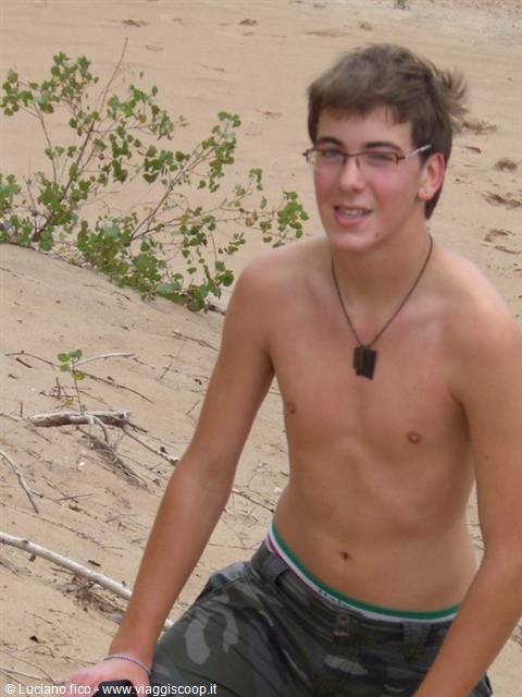 Shirtless boy with glasses and black pendant, sitting on sand