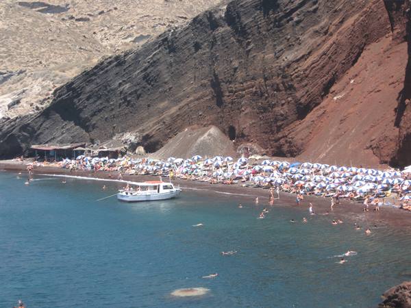 Swimmers and umbrellas on red beach, anchored boat, volcanic cliffs