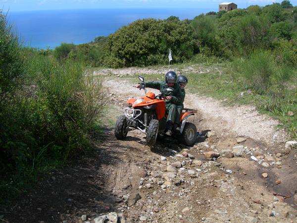 Two people riding an orange quad bike on rocky trail, sea and greenery behind