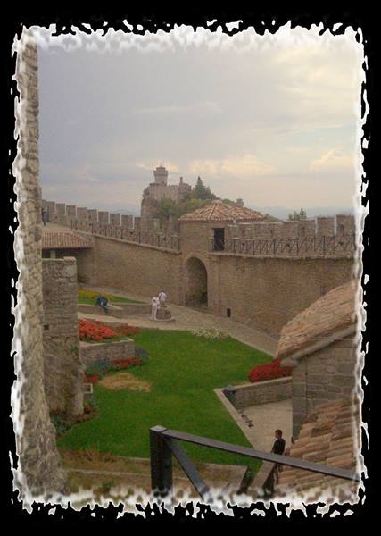 Stone crenellated walls with tower, flowerbeds and tourists under a grey sky