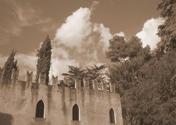 Crenellated castle wall with arched windows, cypresses and clouds, sepia tone