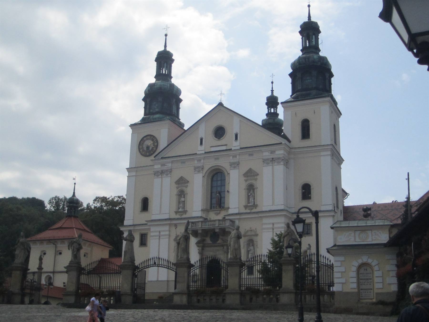 Baroque basilica with two green towers and iron gate, people walking