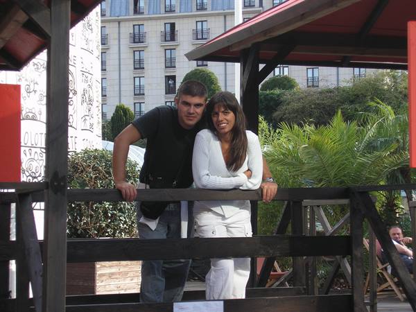 Couple leaning on wooden fence under a pavilion, building and palms behind
