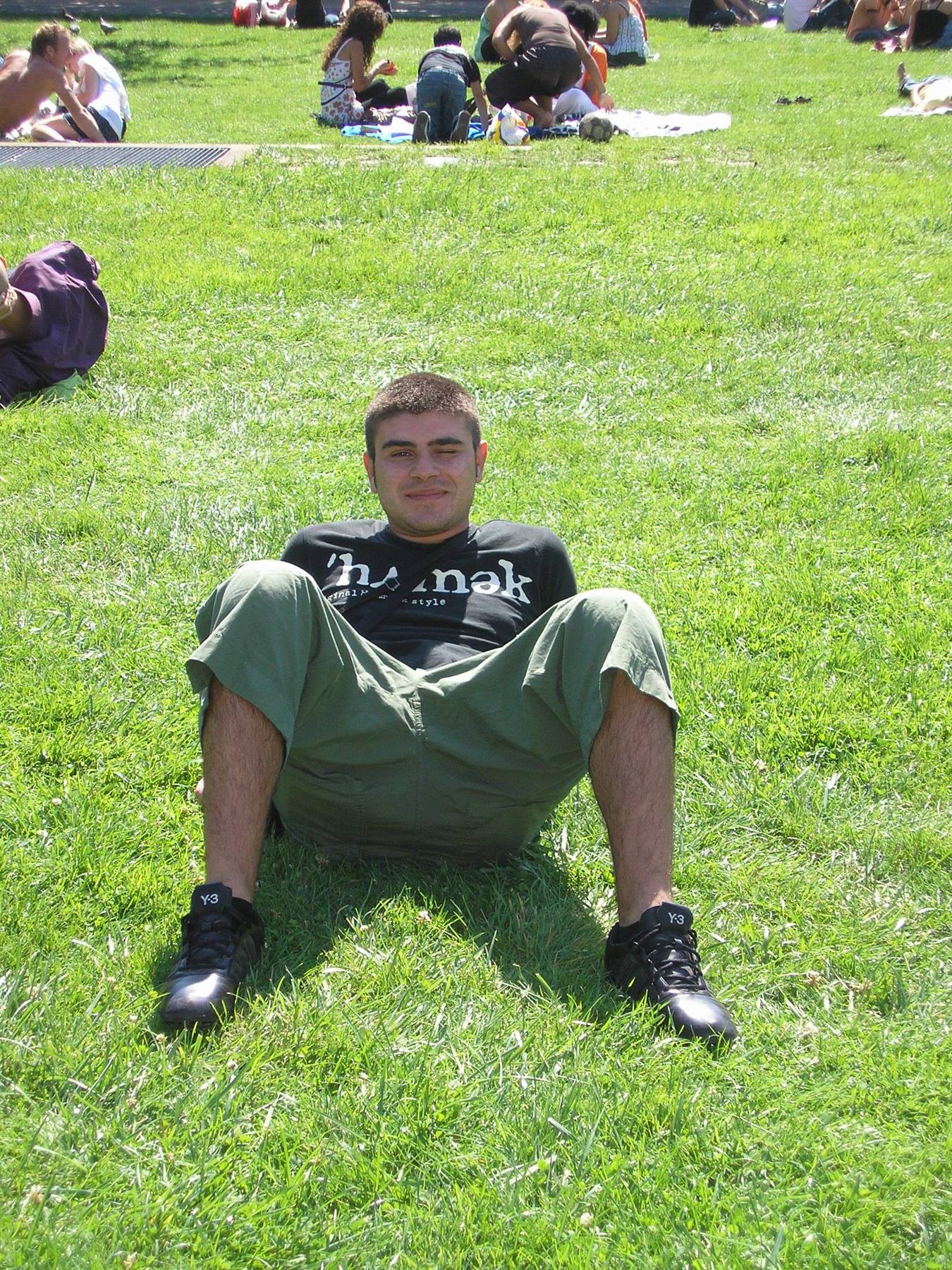 Guy lounging on park grass, people sitting in the sun behind