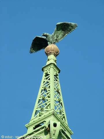 Green bronze eagle with spread wings atop wrought-iron spire against blue sky