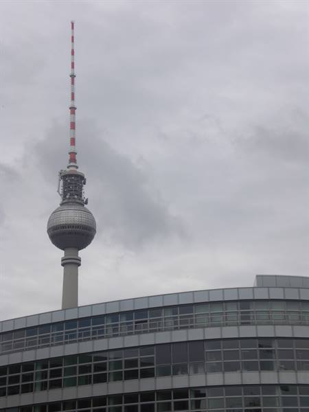 Berlin's Fernsehturm tower rises above a curved glass building under grey sky