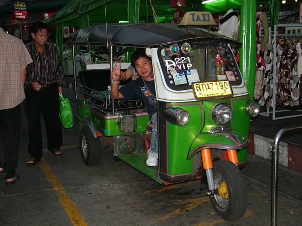 Smiling man gives peace sign on green tuk-tuk parked at night market