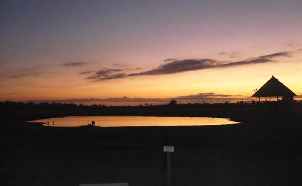 Waterhole reflecting the orange dusk sky, thatched-roof lodge silhouette on the right