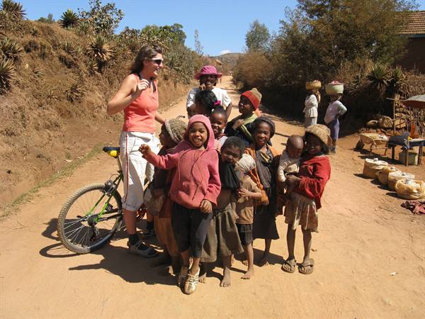 Woman with bicycle surrounded by smiling children on a rural dirt road