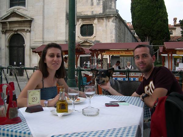 Couple at outdoor café table with dog nearby, market stalls and stone church