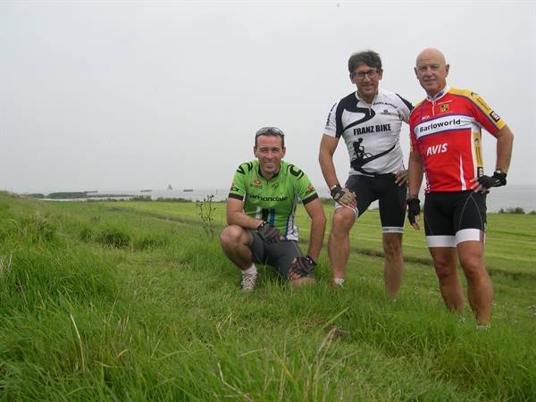 Three cyclists in cycling jerseys pose on grassy dike under grey sky