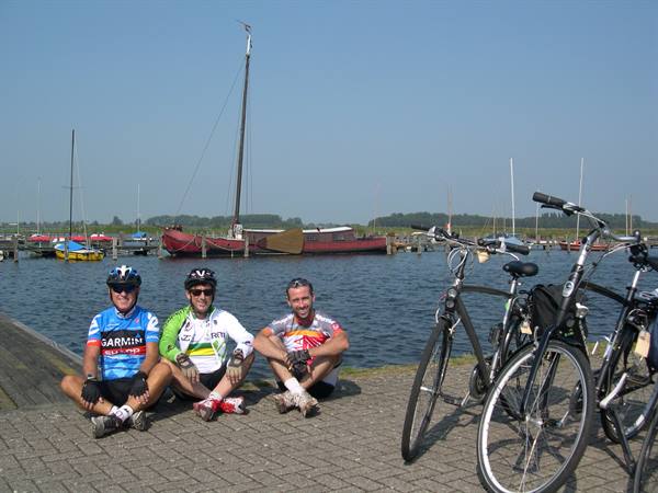 Three cyclists sitting on the dock beside their bikes, sailboats behind