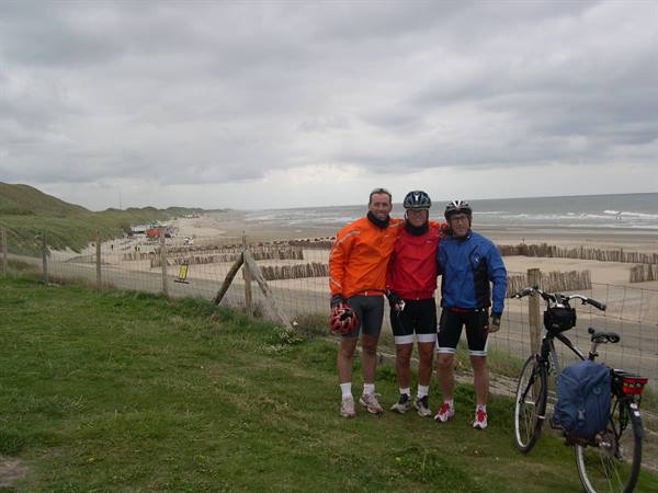 Three cyclists in windbreakers beside a loaded touring bike, facing a windy beach with wooden fences