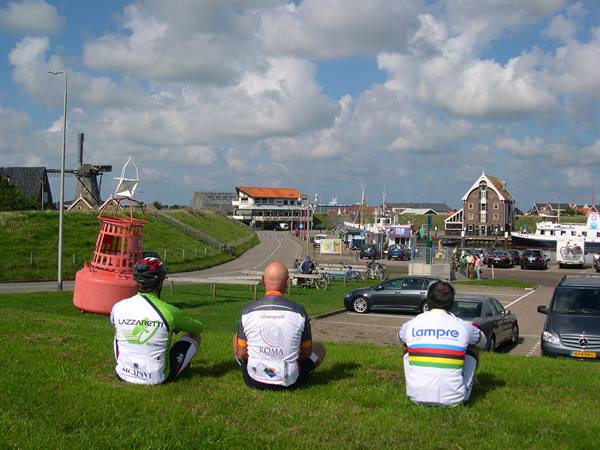 Three cyclists in jerseys sitting on grass facing a harbor and a windmill