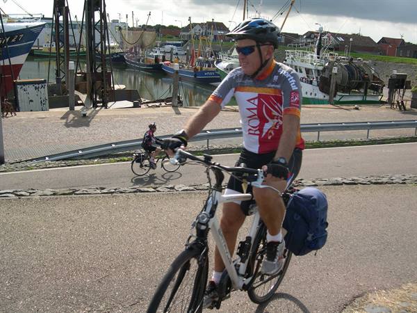 Helmeted cyclist with blue pannier rides past fishing boats in harbor