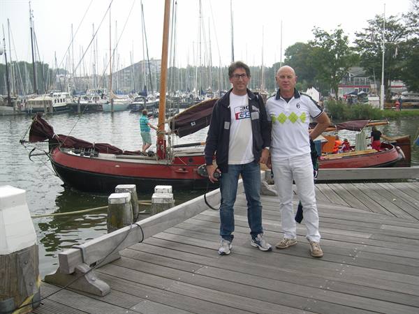 Two smiling men on wooden dock, sailboats moored in the harbor