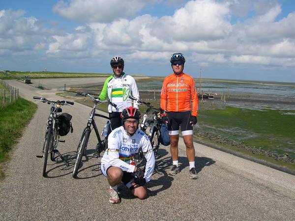 Three cyclists with helmets beside their bikes on a dike, mudflats and clouds