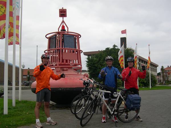 Three cyclists pose with their bicycles beside a large red buoy, grey sky