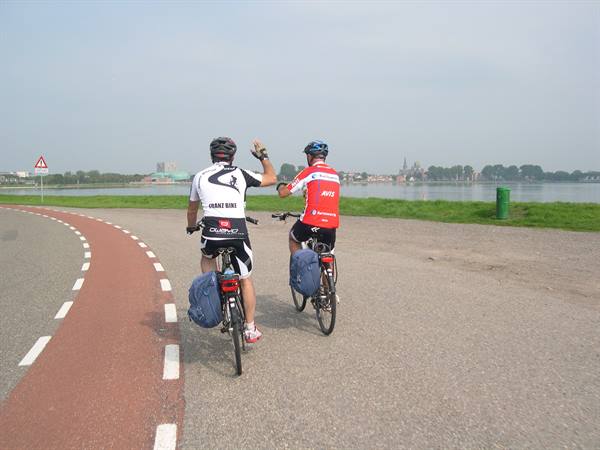 Two touring cyclists wave while riding on a paved bike path by the lake