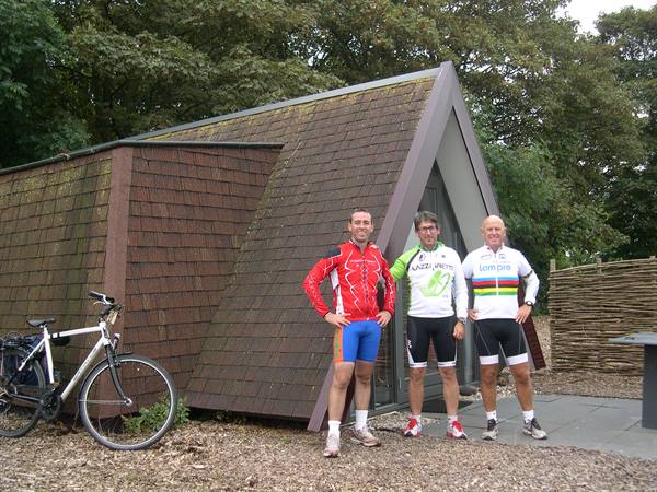 Three cyclists in sport jerseys next to a bicycle in front of a wooden cabin