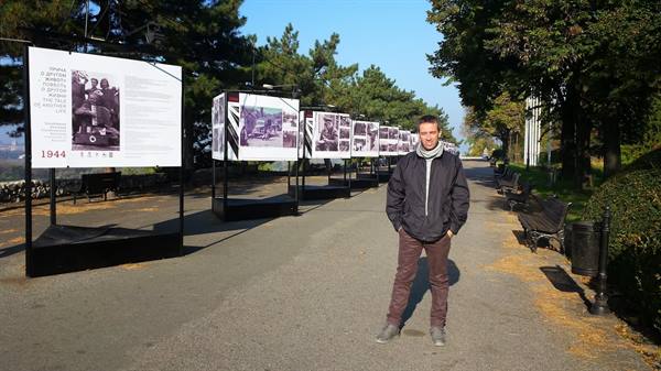 Man in black jacket beside outdoor photo exhibition panels along a tree-lined path