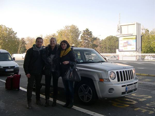 Three friends beside a grey Jeep with red suitcases on a tree-lined street