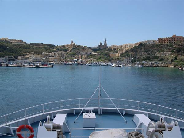 Ferry bow approaching Gozo harbour, moored boats and church spires on the hillside