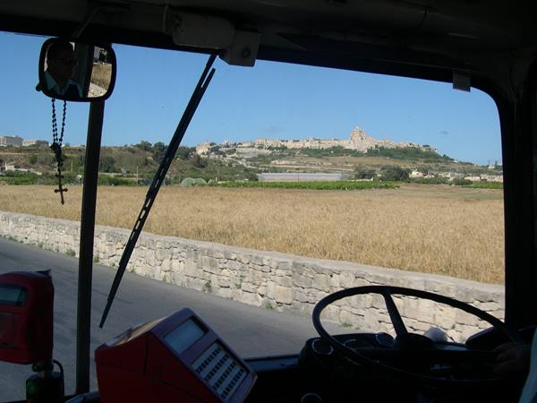 Inside a Maltese bus: steering wheel, rosary and ticket machine, fields toward Mdina