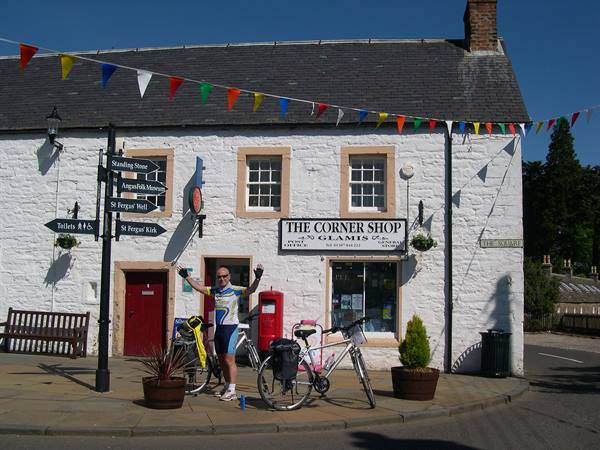 Cyclist with raised arms outside the Corner Shop in Glamis, whitewashed building and flags