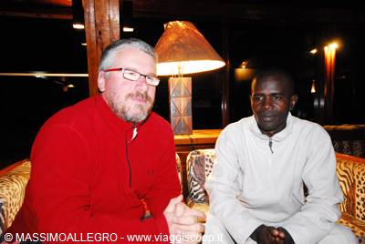 Two men sitting on a leopard-print couch beside a glowing wooden lamp