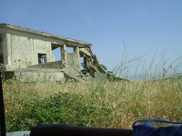 Ruined concrete building amid dry grass, photographed from the car under blue sky