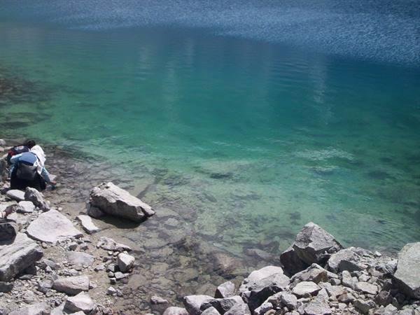 Hiker with backpack crouches by turquoise water among grey boulders on lake shore