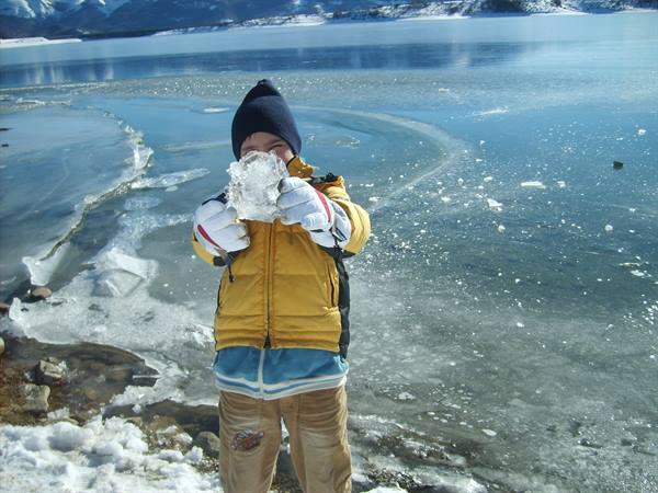 Child in yellow jacket and blue hat holds an ice chunk on frozen lake