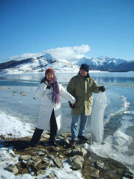 Couple on frozen lakeshore, man lifting ice sheet, snowy mountains behind
